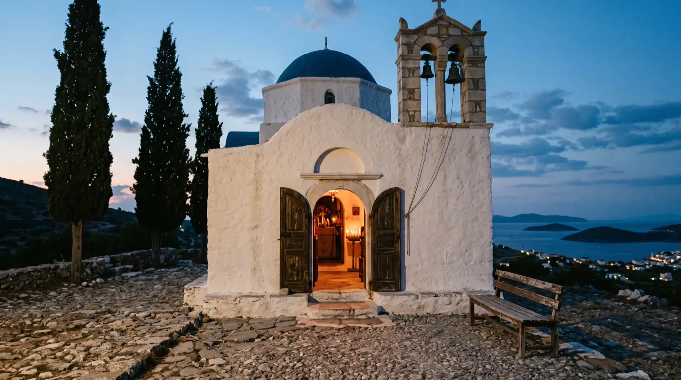 A whitewashed chapel with a blue dome and bell tower at dusk, its door open to a candlelit interior above an island bay