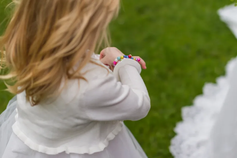 Young child in a white dress and cardigan wearing a colorful beaded bracelet, standing on green grass beside a bride’s lace gown.