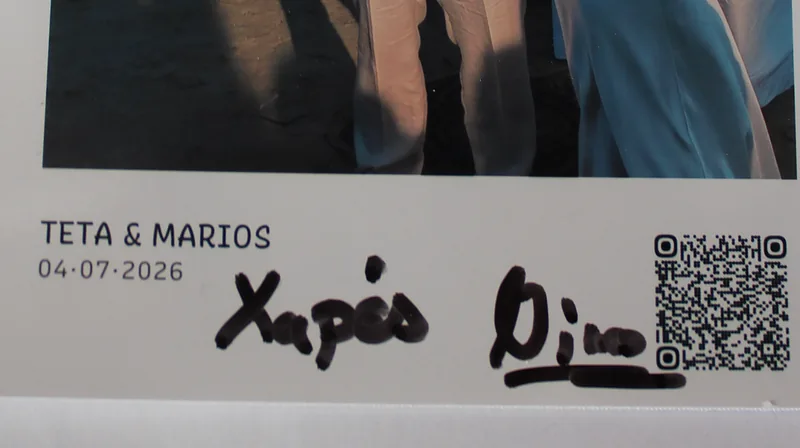 Close-up of a printed card's caption strip reading Teta and Marios 04-07-2026 next to a QR code, with a guest's handwritten wish in black marker across the white border
