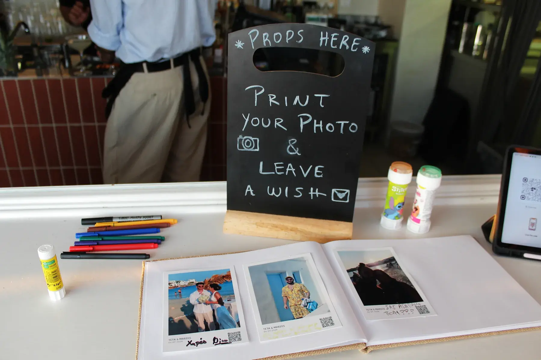 A Smart Print station at a Greek island wedding, with a chalkboard sign reading Print your photo and leave a wish, next to an open guest book with printed photo cards and coloured pens