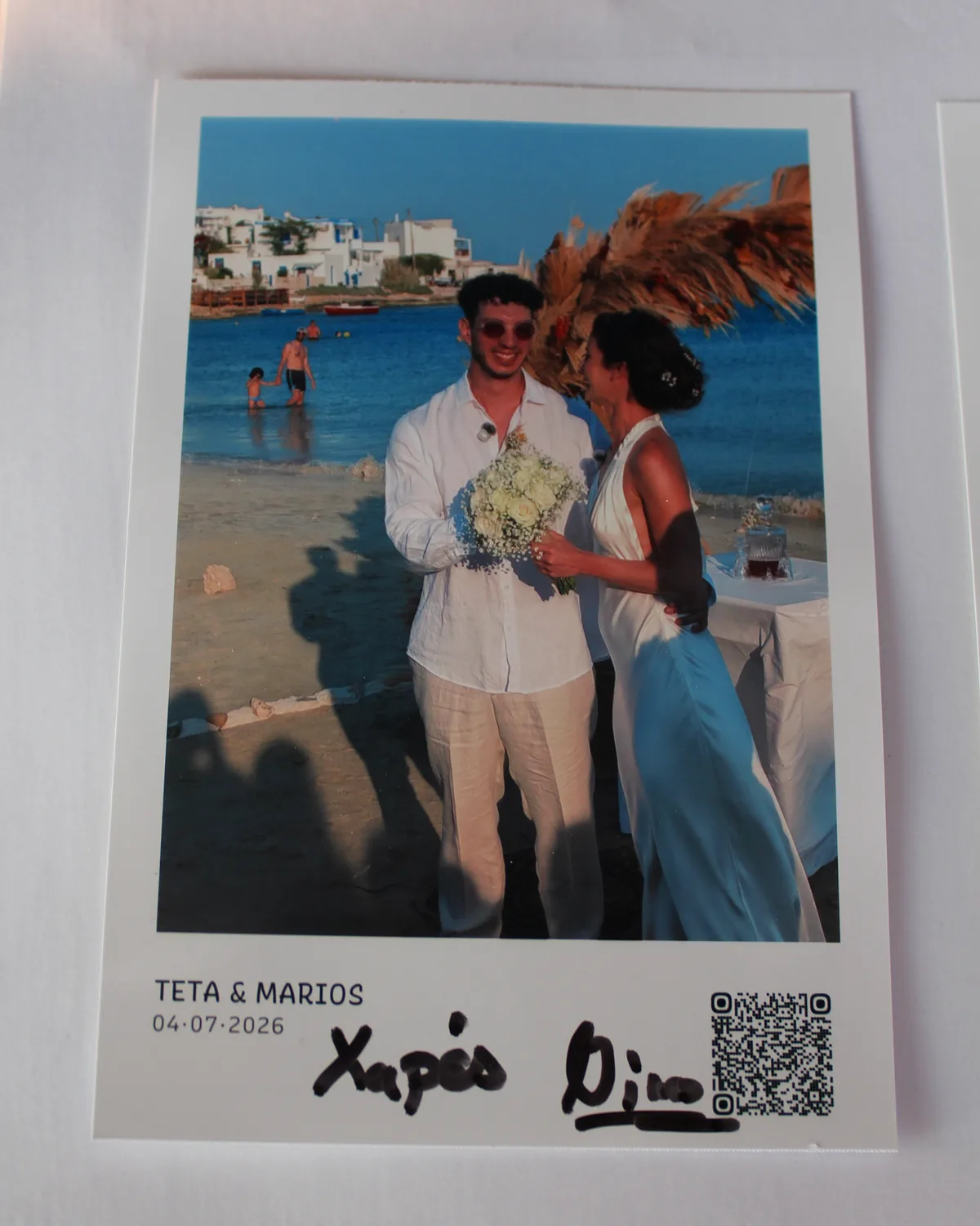 A close-up of a printed Smart Print card showing a guest photo, the caption Teta and Marios 04-07-2026, a QR code, and a handwritten wish in marker pen
