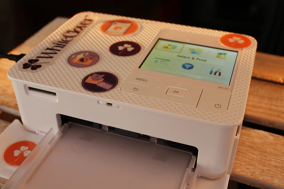 The WhiteClover Smart Print station, a compact dye-sublimation photo printer with WhiteClover branding and clover stickers, printing 4×6 cards at a wedding venue