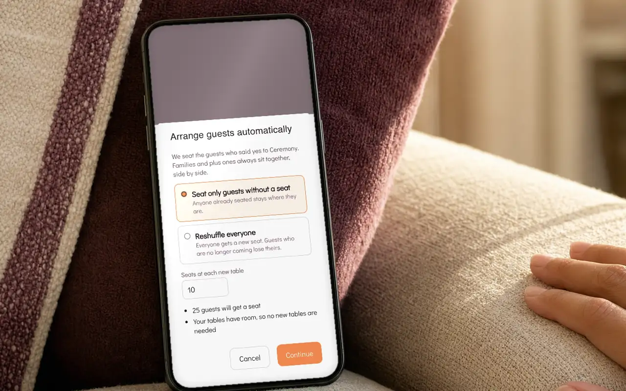 A phone propped against a sofa cushion, showing the Arrange guests automatically dialog with two choices: seat only guests without a seat, or reshuffle everyone