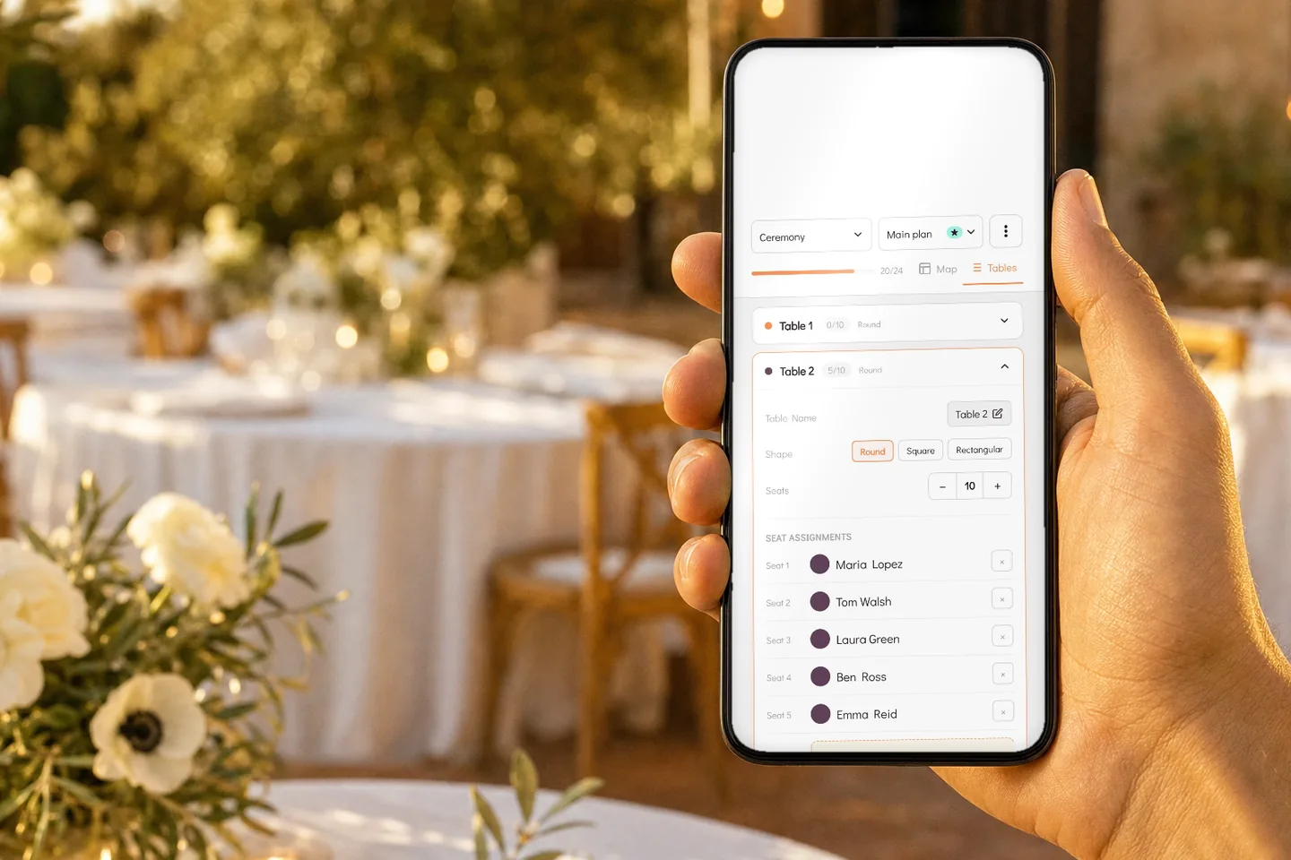 A hand holding a phone above a reception table set outdoors, the screen showing the WhiteClover seating chart with Table 2 and its five seated guests