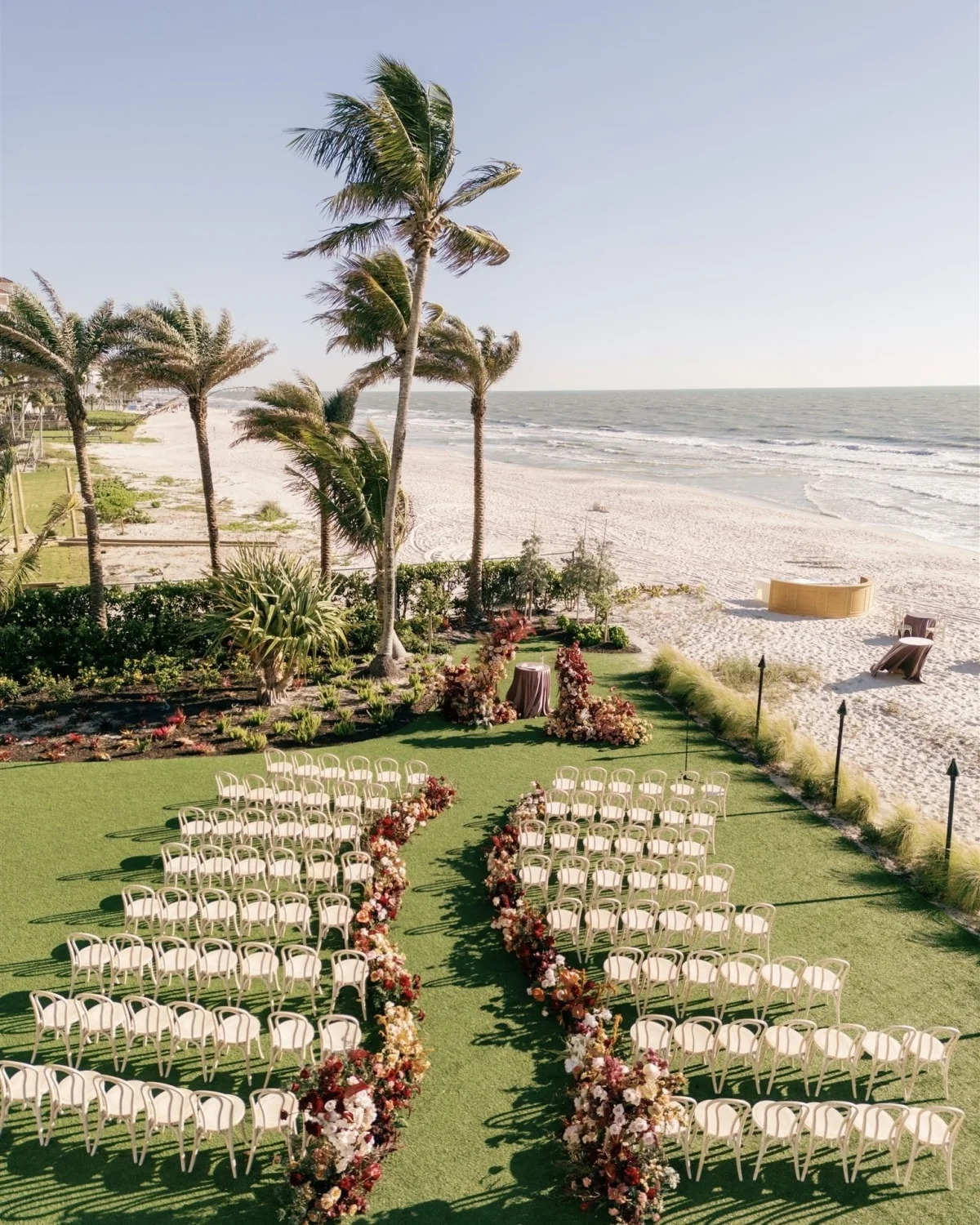Τελετή γάμου της A. Marie Events & Design δίπλα στον ωκεανό, με λευκές καρέκλες και διάδρομο από κόκκινα και ροζ λουλούδια