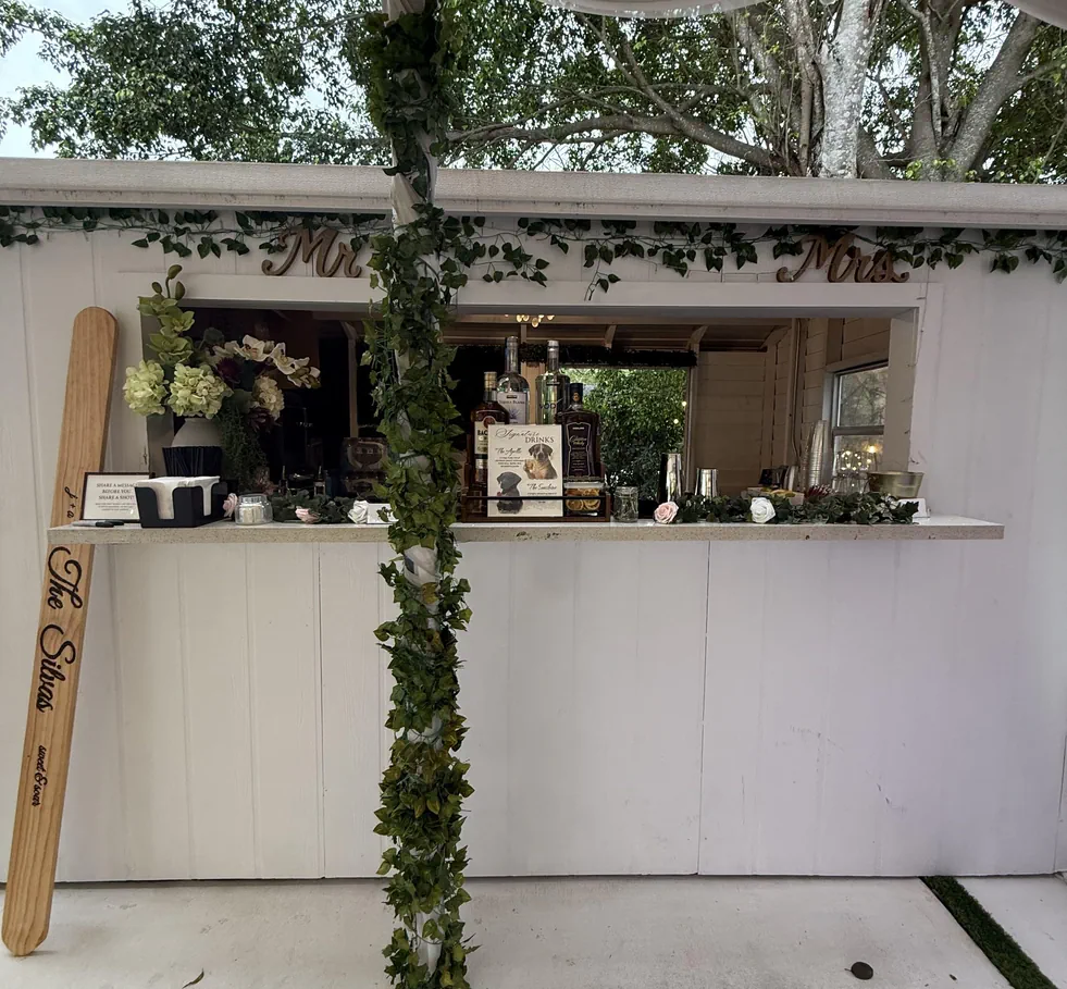 Mobile wedding bar by Sip and Dips featuring a white wooden setup with greenery garlands, floral décor, and displayed spirits, serving drinks in an outdoor event setting.