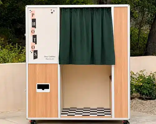 Enclosed wedding photo booth with wood panel exterior, green curtain entrance, and black-and-white checkered floor, set outdoors for private guest photo sessions.