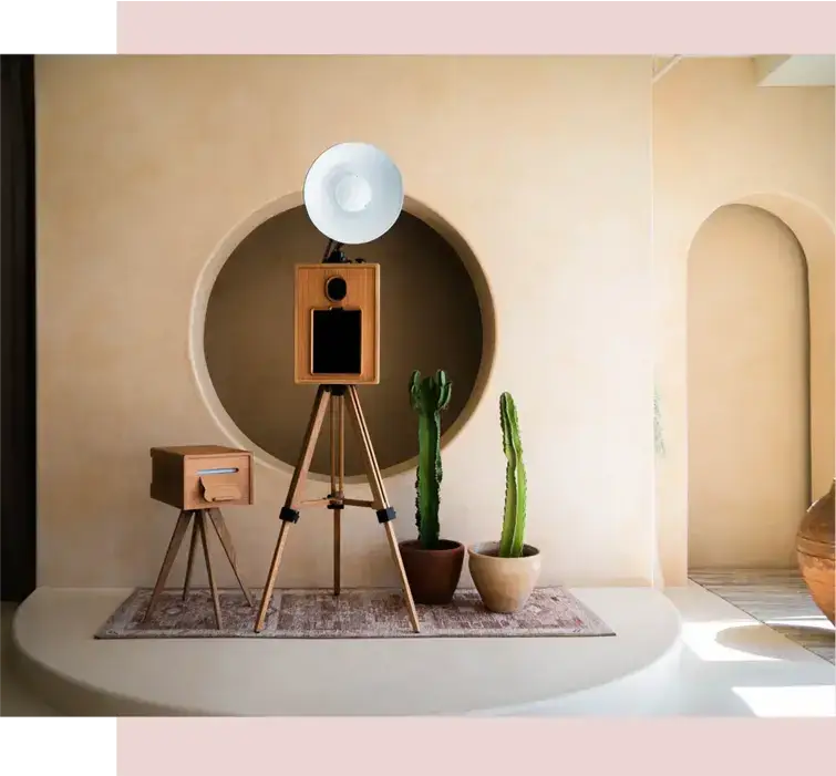 Vintage-style photo booth camera on a wooden tripod with flash reflector, set in a minimalist wedding venue with beige arches, neutral tones, and potted cacti decor.
