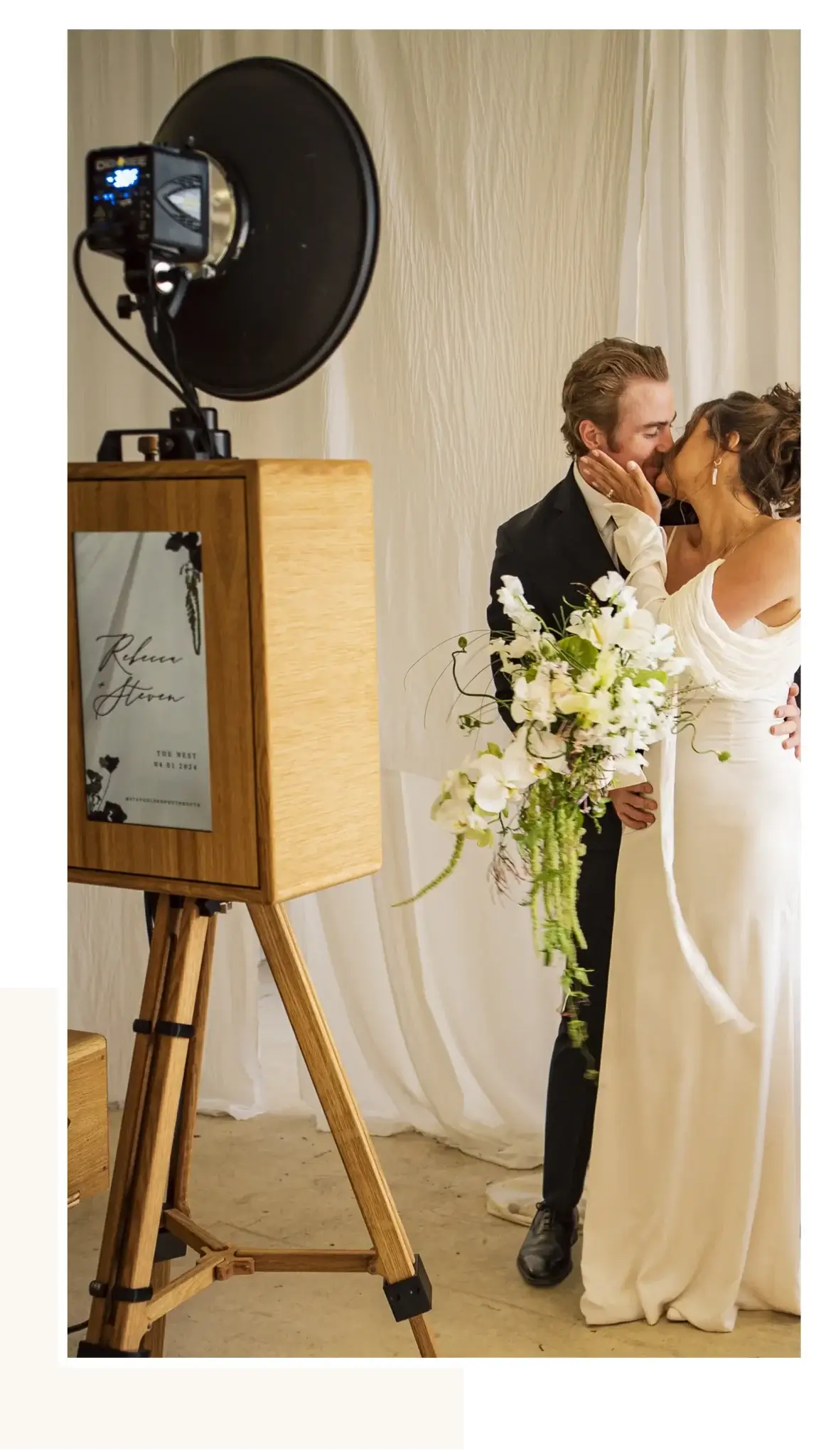 Vintage wooden wedding photo booth with studio flash on tripod capturing a couple kissing, one holding a cascading white floral bouquet, set against soft draped fabric backdrop.