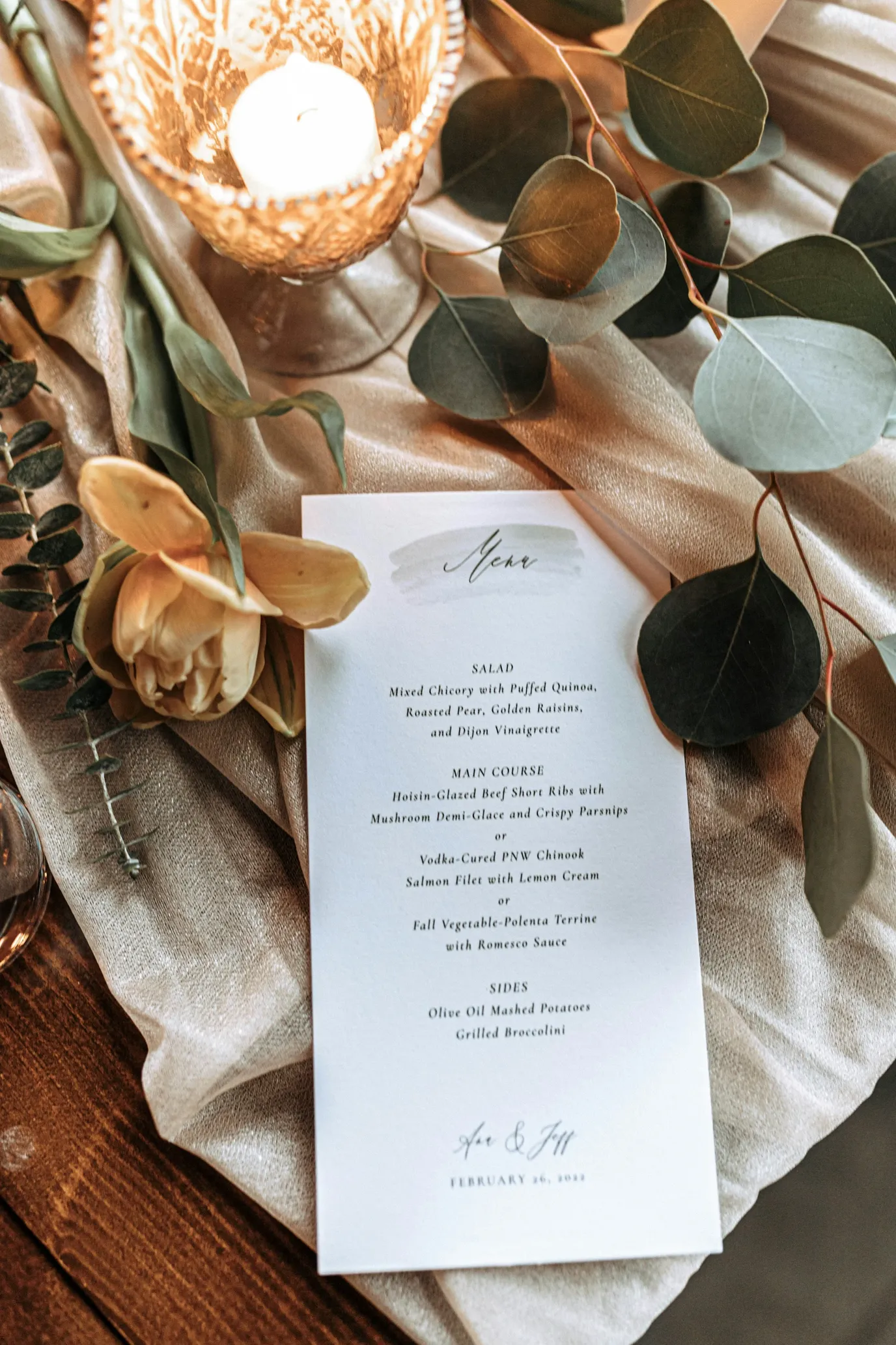 Elegant wedding menu card placed on a satin table runner, surrounded by eucalyptus leaves, a candle in a glass holder, and a soft yellow flower.