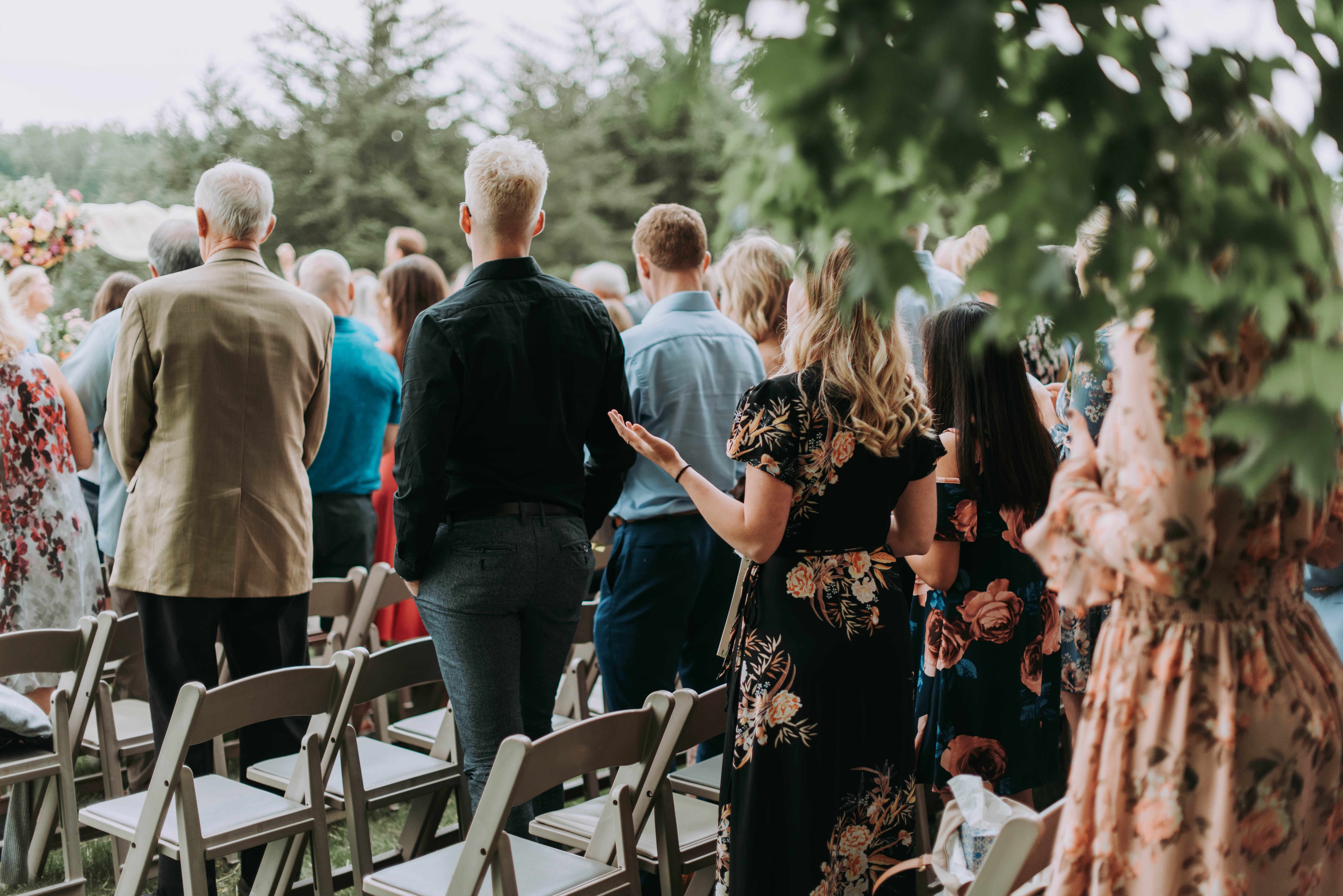 Guests at an outdoor wedding ceremony, standing and facing forward, with their attention directed towards the event, showcasing a variety of elegant attire in a serene, tree-lined setting.